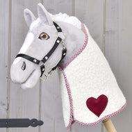 Plyšový Hobby Horse od Hobby Horse LarDen s bílošedým obličejem, černou uzdečkou a fleecovou Dekou Cube Red s červenou nášivkou ve tvaru srdce, nasazený na dřevěné tyči na pozadí světlého dřeva.
