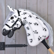 Hobby Horse Chantal s černou ohlávkou od Hobby Horse LarDen, s bílou srstí s černými skvrnami, šedou přízovou hřívou, černou ohlávkou a na dřevěné tyči, před světle šedou dřevěnou stěnou.