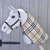 Hobby Horse LarDen „Deka Cremo England“ na šedém Hobby Horse s černou ohlávkou a béžovým kostkovaným kabátkem, doplněná čabrakou pro ochranu před hmyzem, nasazeném na dřevěné tyči před šedou dřevěnou stěnou.