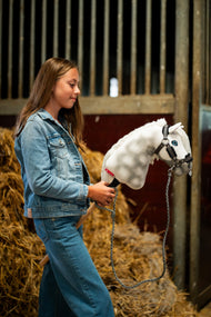 Dívka v džínové bundě stojí před balíky slámy ve stáji a drží Hobby Horse Elektra s černou ohlávkou od Hobby Horse LarDen, bíle grošovaná plyšová hlava, modré oko, bílá přízová hříva, s ohlávkou Graphite Full s kožešinou z koženky a pleteným vodítkem