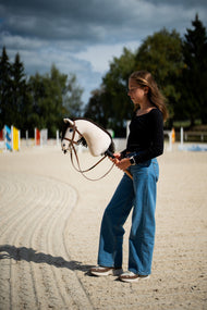 Dívka v černém topu stojí na pískové jízdárně pod bouřkovou oblohou a drží Hobby Horse Frederik LD s černou ohlávkou od Hobby Horse LarDen, v pozadí jsou barevné skokové tyče, krémová plyšová hlava, tmavě hnědá přízová hříva, s kompletní hnědou uzdečkou s nánosníkem