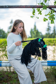 Dívka ve světlém svetru sedí na hrazení výběhu a sbírá otěže Hobby Horse Maximus s černou ohlávkou od LarDen, s černou plyšovou hlavou a dlouhou černou přízovou hřívou, s černou koženkovou ohlávkou se zeleným kožíškem a černými koženkovými otěžemi