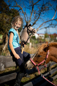 Blond dívka v tyrkysovém tričku sedí na dřevěném výběhovém plotu a drží Hobby Horse Piccolino s černou Ohlávkou od Hobby Horse LarDen se světle béžovou plyšovou hlavou a tmavou hřívou z příze, za ní stojí ryzý poník, s kompletní černou koženkovou uzdečkou