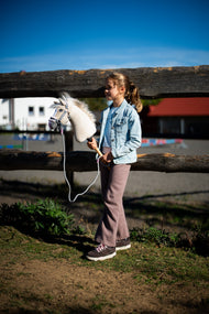 Lifestyle photo: a girl in a light denim jacket and mauve trousers stands at a wooden paddock fence holding the Hobby Horse Pixie with Black Halter by Hobby Horse LarDen - beige plush head with a white yarn mane - shown here with the Halter grey with pink fur and the rope stripe Tizian lead.