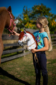 Dívka v tyrkysovém tričku drží Hobby Horse Rufus s černou Ohlávkou od Hobby Horse LarDen s bílou plyšovou hlavou se skvrnami Appaloosa a červenou hřívou z příze vedle ryzého poníka ve slunném výběhu, s bordó koženkovou sadou s uzdečkou, otěžemi a martingalem