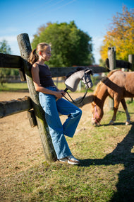 Dívka v hnědém topu a džínách se opírá o dřevěný sloupek plotu a drží Hobby Horse Thor s Black Ohlávkou od Hobby Horse LarDen, šedou plyšovou hlavu s černou přízovou hřívou, za ní se na ohradě pase poník, s černou uzdečkou Complete s diamanty