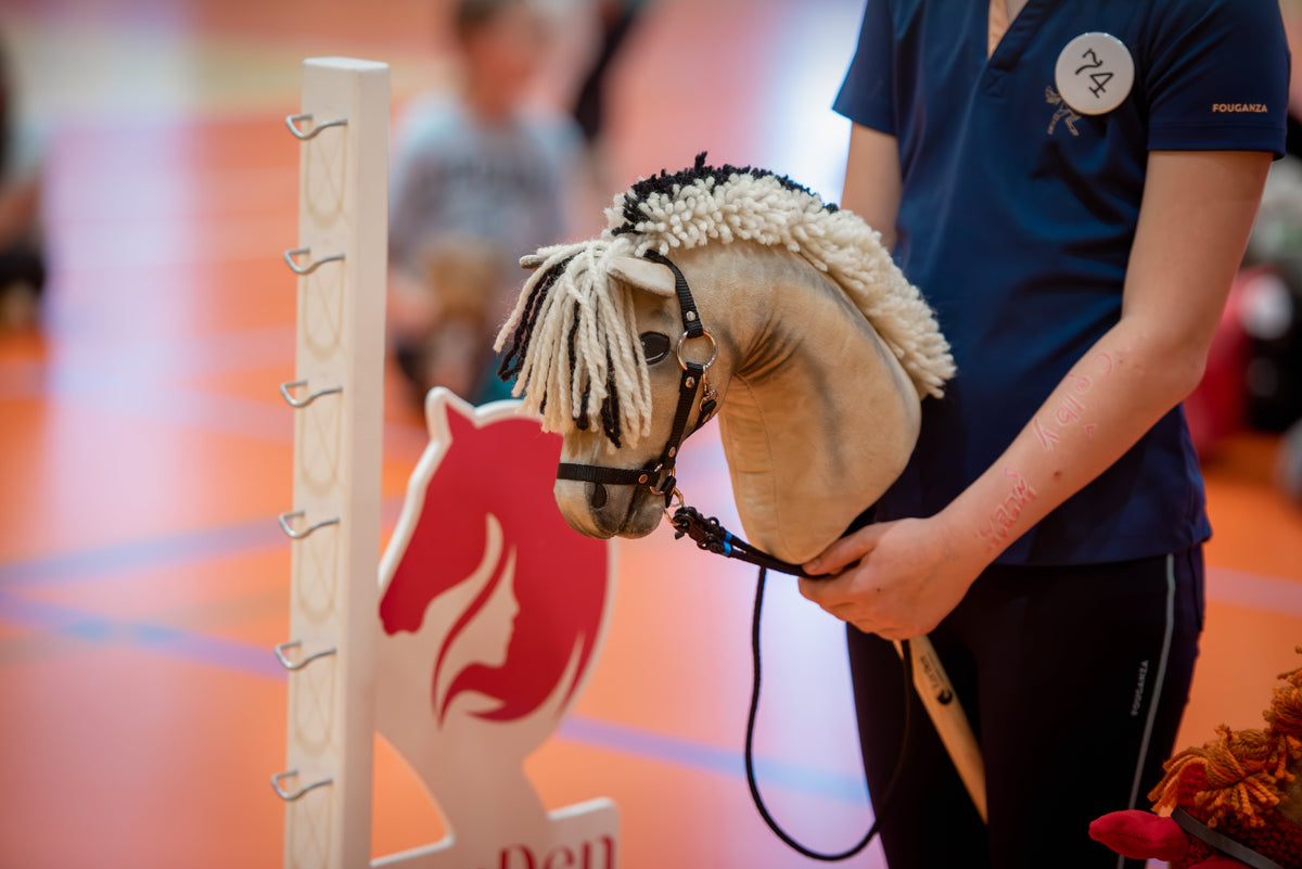 Detail z halových závodů LarDen a CHHA, kde účastníci v tmavě modrých sportovních tričkách drží Hobby Horse vedle bílé parkurové stojky a červeno-bílého křídla skoku; popředí vyplňuje světle hnědý Hobby Horse s krémovo-černou přízovou hřívou a uzdečkou.