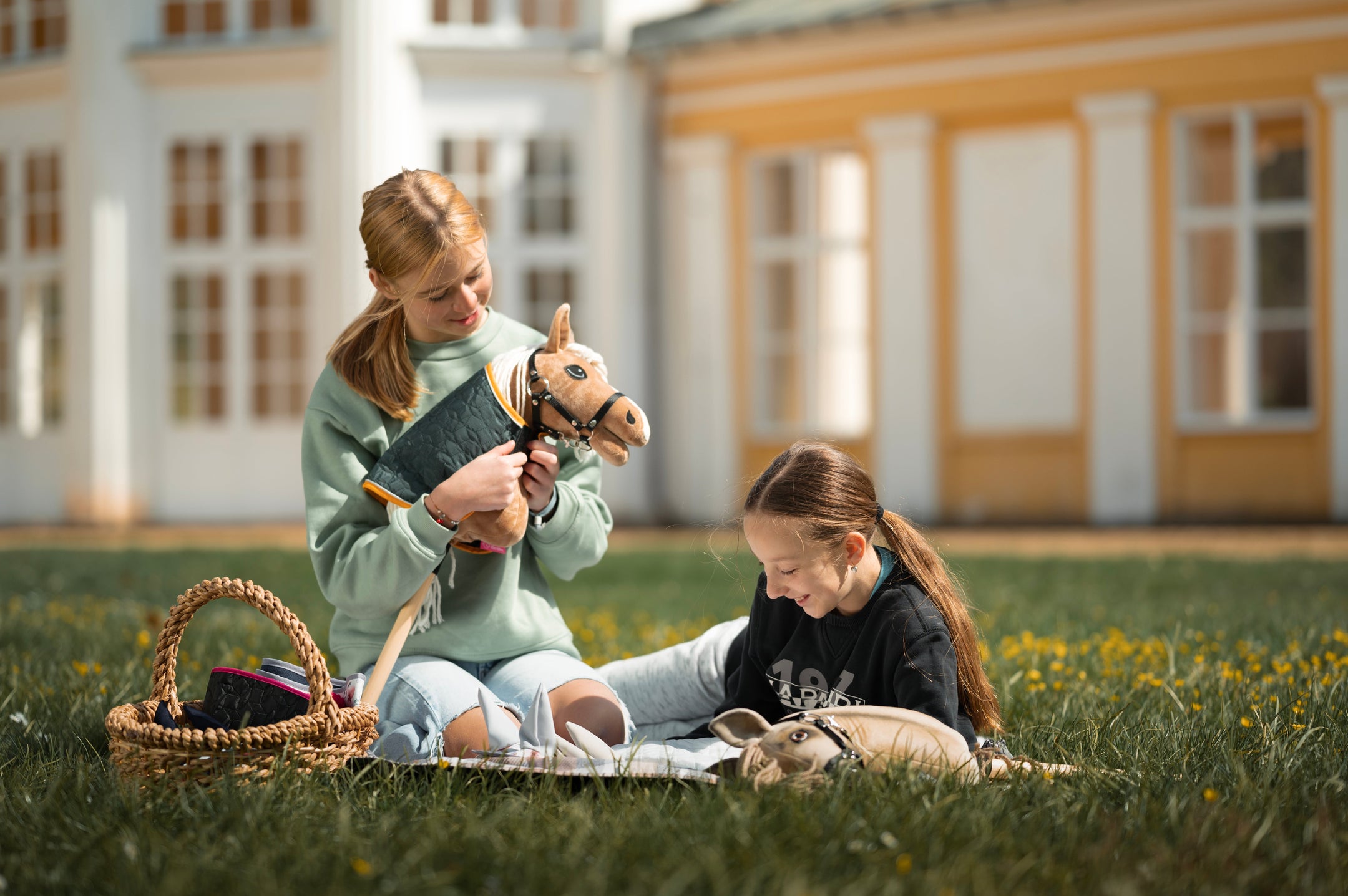 A girl in a pale green sweatshirt sits on the grass beside a wicker basket of hobby horse gear, gently holding a tan hobby horse with a black bridle, white mane, and quilted cover. Another child is partly visible on a blanket nearby, creating a calm