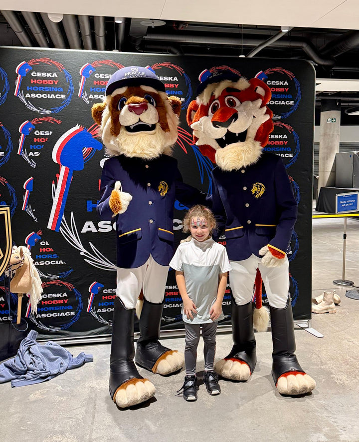 A smiling young girl with face paint stands between two Prague Playoffs animal mascots in navy riding-style jackets and caps, posing in front of a Česká Hobby Horsing Asociace backdrop in the O2 Arena concourse. A small hobby horse rests at the side