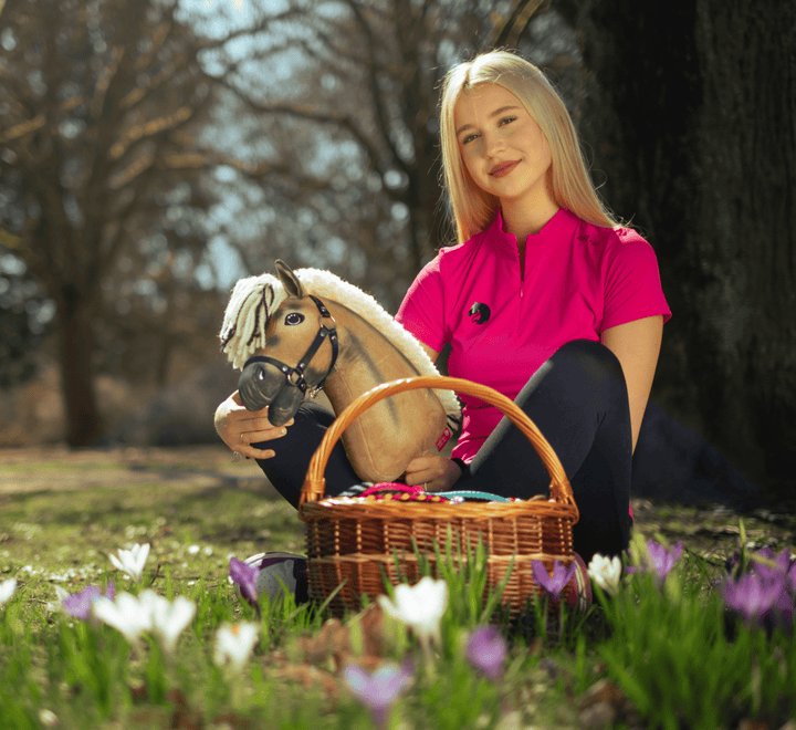 Mladá žena v jasně růžovém tričku sedí venku na trávě, drží hračku Hobby Horse a proutěný košík plný barevných vajec, obklopená rozkvetlými bílými a fialovými květy.