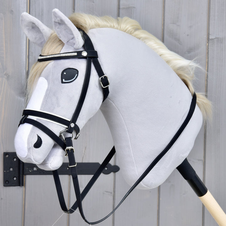 Plyšový Hobby Horse s bílou hřívou, černou uzdečkou a bílým pruhem na obličeji je připevněný k dřevěné tyči a je umístěný před šedým dřevěným pozadím.
