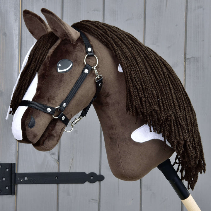 Realistický hnědobílý Hobby Horse s černou uzdečkou a dlouhou hřívou z příze, nasazený na dřevěné tyči, umístěný před šedým dřevěným plotem.
