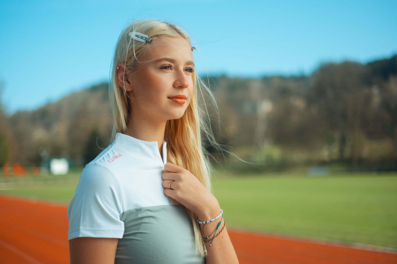 Mladá blond žena ve sportovním oblečení stojí na venkovní běžecké dráze a zamyšleně drží svůj hobby horse. V pozadí je zelená tráva, stromy a modrá obloha, což vystihuje její vášeň pro Hobby Horsing.