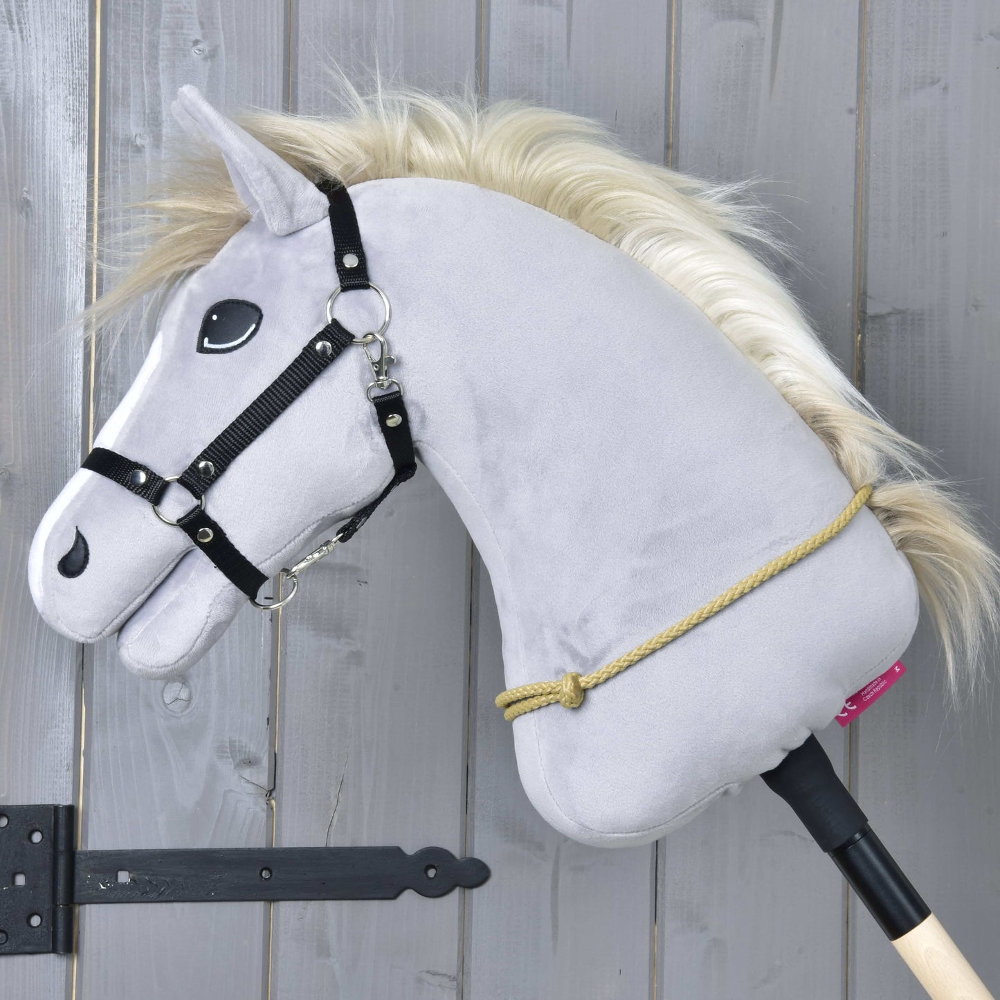 Šedý Hobby Horse LarDen s černou uzdečkou, zlatou hřívou a ručně vyrobeným Nákrčníkem Nature visí na dřevěné tyči před šedou stěnou. Kolem krku má omotané žluté polypropylenové lano.