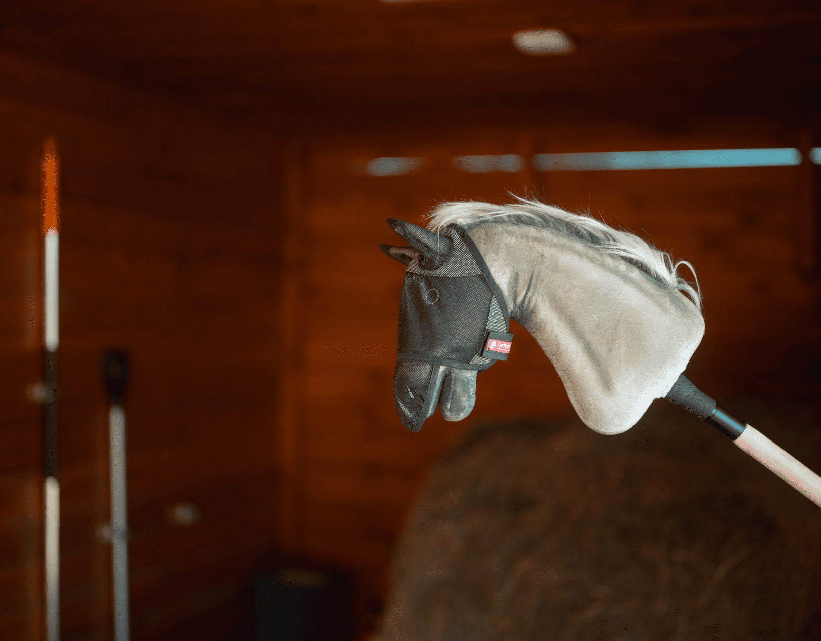 Hobby Horse se šedou plyšovou hlavou a černou uzdečkou je nasazený na tyči uvnitř dřevěné stodoly, obklopený farmářským nářadím a senem – ideální pro podnícení nápaditých dobrodružství v Hobby Horsing.