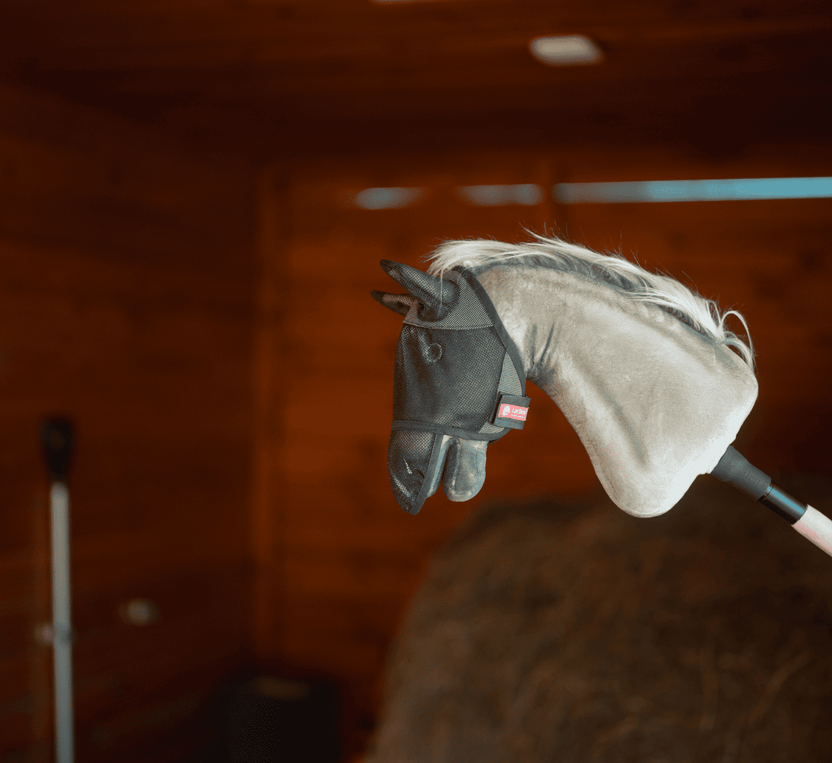 Plyšová hlava Hobby Horse na tyči s černou síťovanou maskou proti hmyzu je vyfocená v interiéru s dřevěnými stěnami a balíkem sena – ideální pro nápaditou zábavu s Hobby Horsing.
