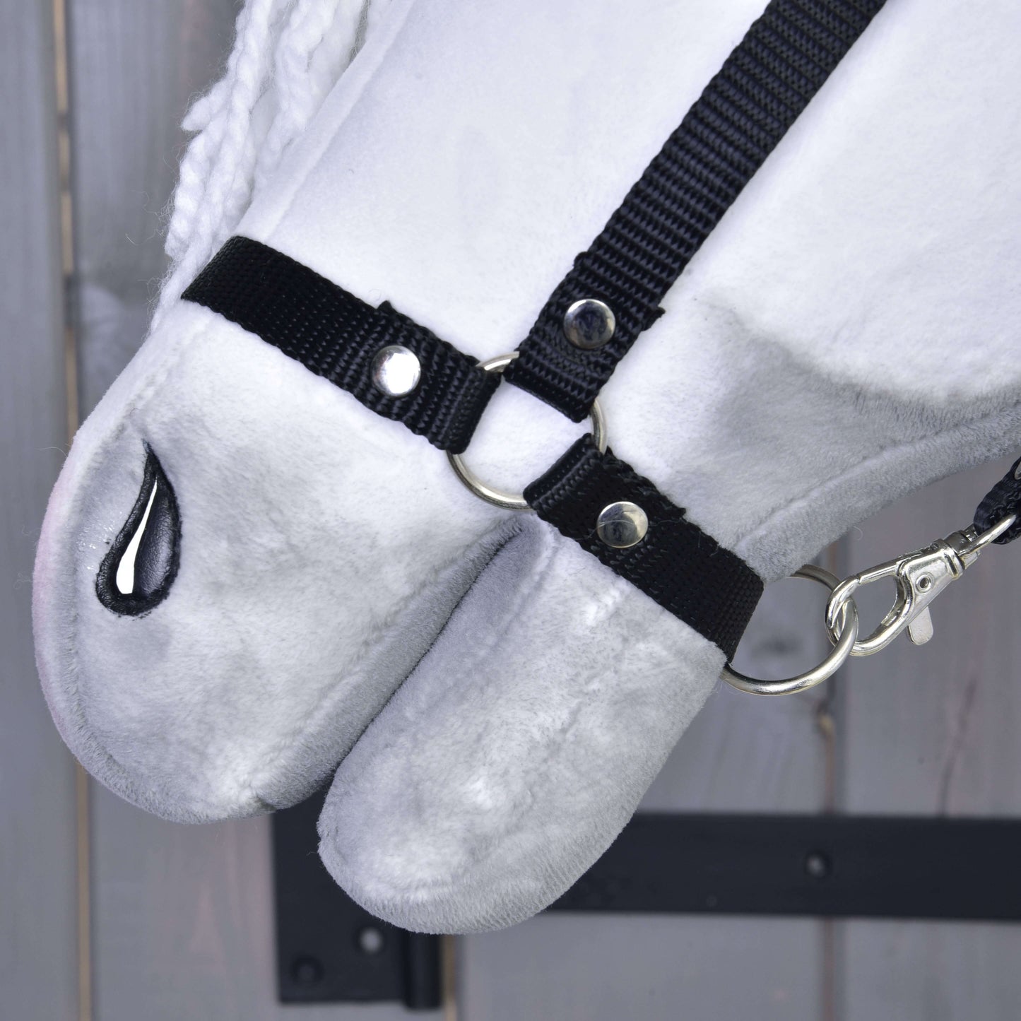 Detail Hobby Horse Elektra s černou ohlávkou od Hobby Horse LarDen s plyšovým bílým obličejem, černou látkovou ohlávkou se stříbrnými kroužky a karabinou a černě prošitou nozdrou, na dřevěném pozadí.