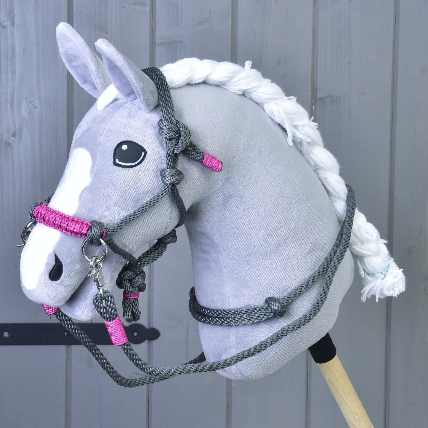 Plyšový šedo-bílý Hobby Horse se zapletenou hřívou a černýma očima předvádí Provazový set Šedá (vel. M) od Hobby Horse LarDen před dřevěným obložením a zvýrazňuje jedinečnou ručně vyráběnou jezdeckou výbavu.