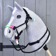 Šedý plyšový Hobby Horse od Hobby Horse LarDen s bílou lysinou a hřívou, s ohlávkou Provazový set Duha, nasazený na dřevěné tyči před pozadím ze šedých dřevěných panelů.