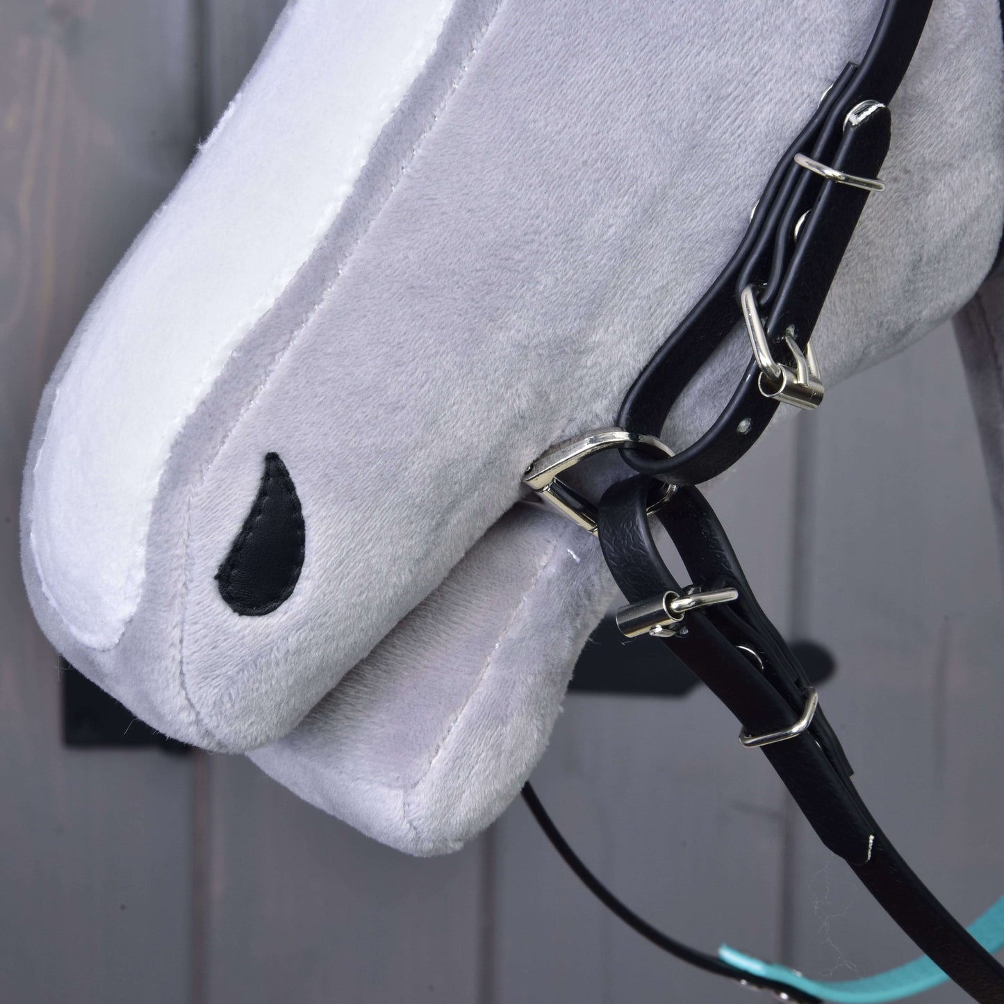 Detail šedého Hobby Horse od Hobby Horse LarDen s Kompletní uzdečkou s tyrkysovou čelenkou bez nánosníku a otěžemi, s černým okem ve tvaru kapky, na rozmazaném šedém pozadí.