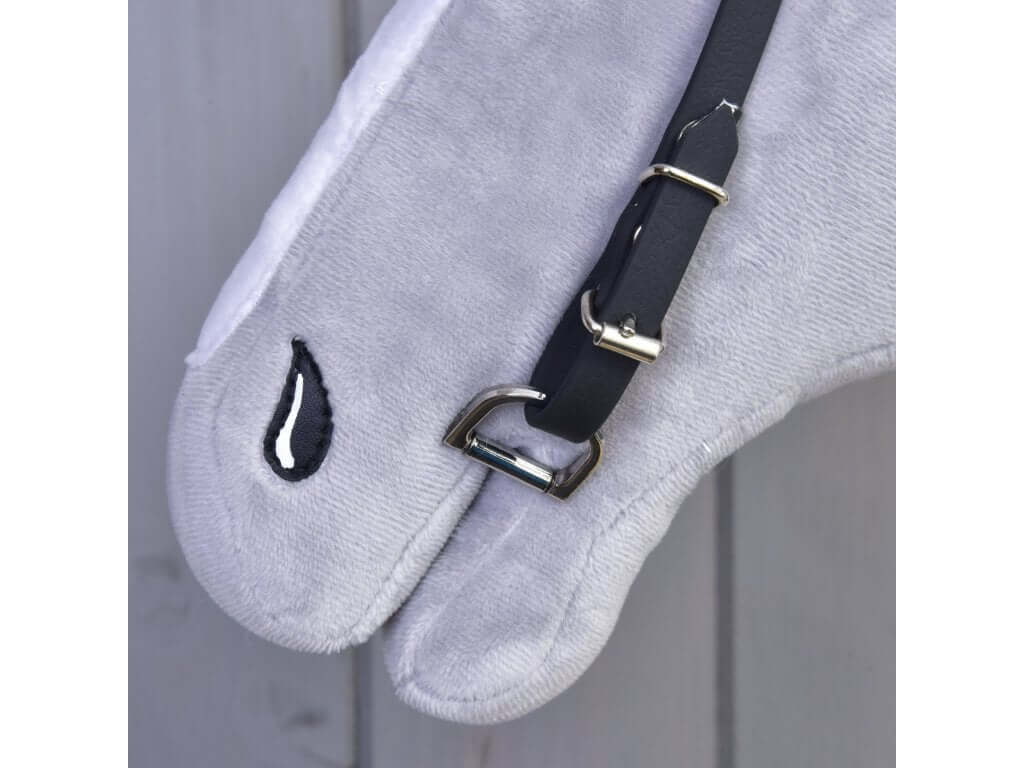 Detail uzdečky Uzdečka s čelenkou bez nánosníku modrá (vel. S) od Hobby Horse LarDen na šedém plyši ve tvaru koňské hlavy, s kovovou přezkou a jednoduchým černobílým vyšívaným okem před světlými dřevěnými panely.