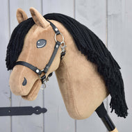 Hobby Horse Samantha s černou ohlávkou od Hobby Horse LarDen má béžovou látkovou hlavu, černou přízovou hřívu a je inspirovaná klisnou anglického plnokrevníka; je zobrazená na dřevěné tyči před šedými dřevěnými panely.