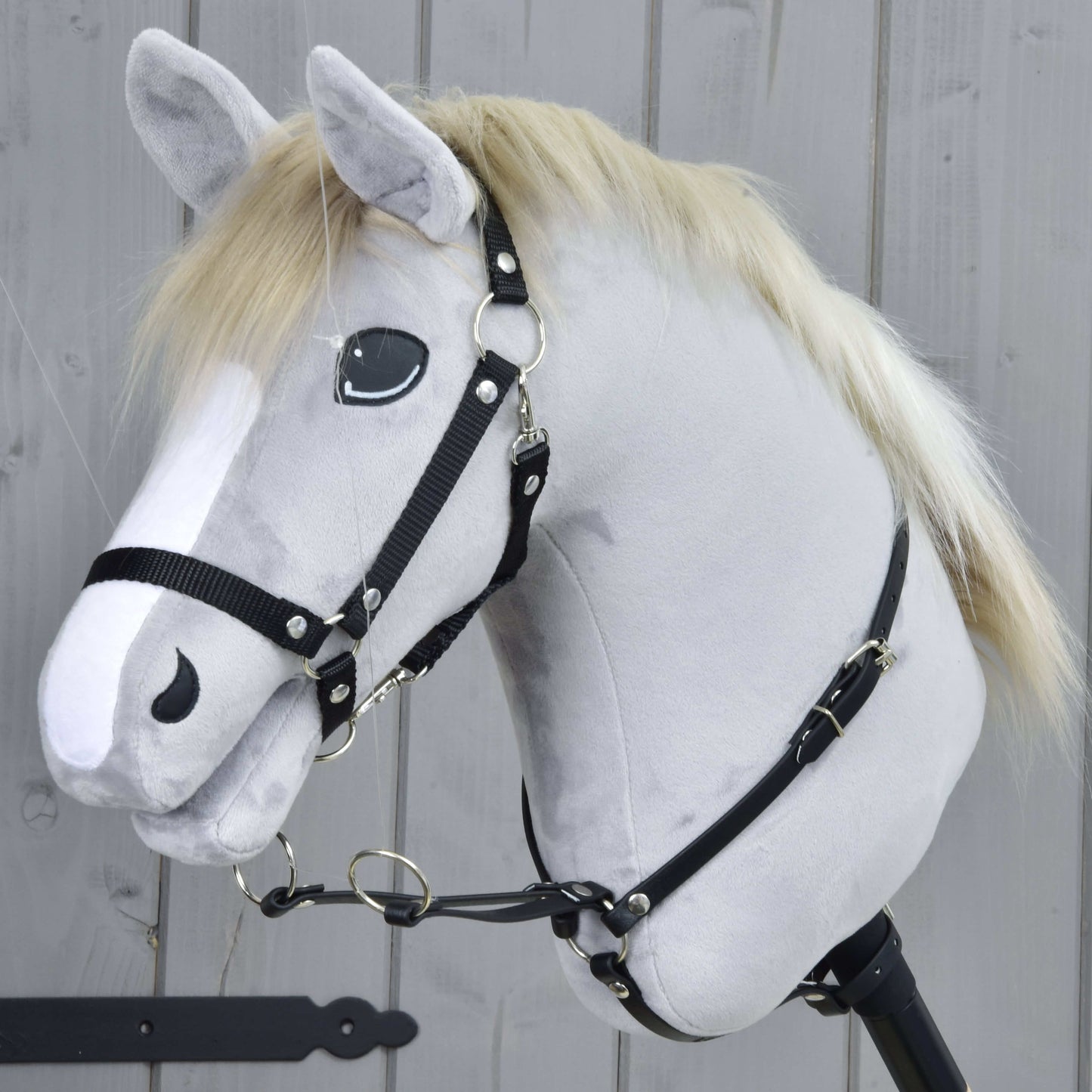 Martingale černý od Hobby Horse LarDen na realistické šedé hračce Hobby Horse se světlou hřívou, bílou lysinou a černou uzdečkou, nasazené na dřevěné tyči před šedým dřevěným plotem.