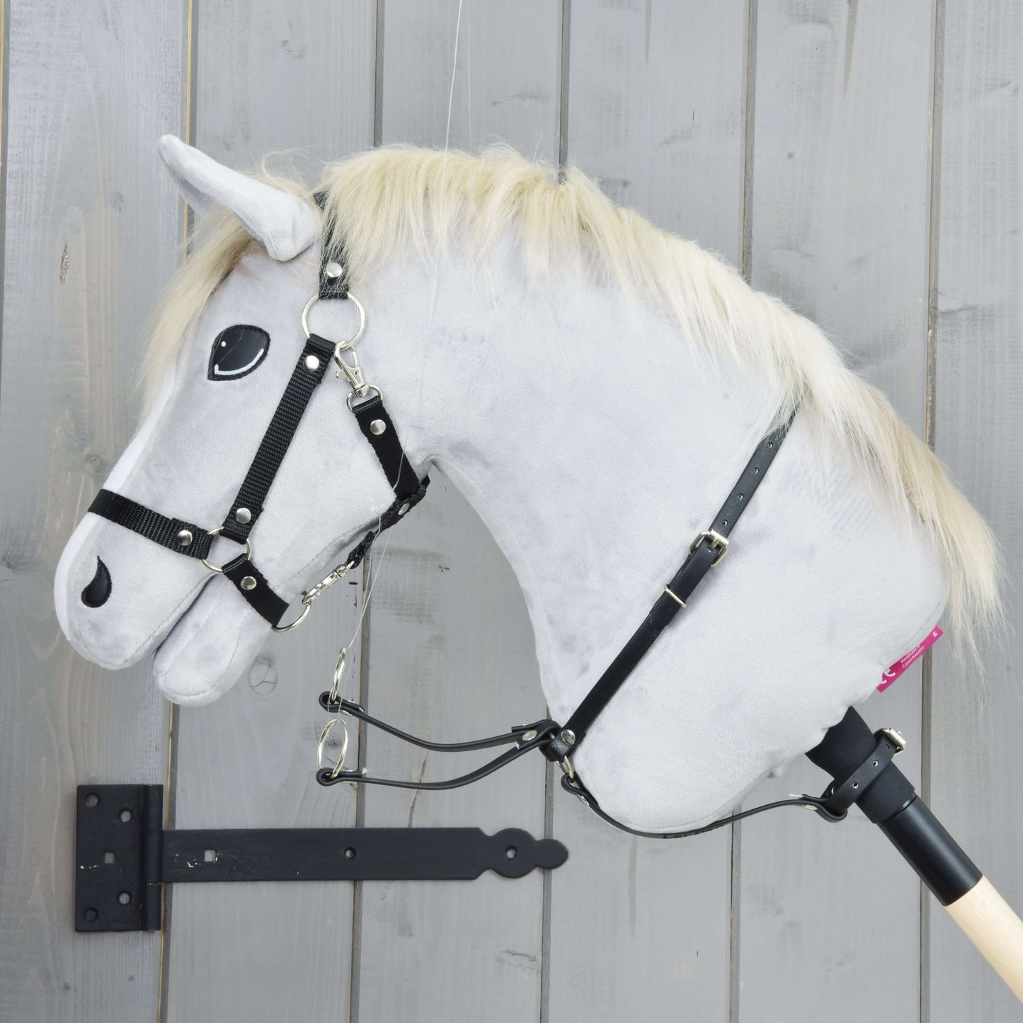 Martingale černý od Hobby Horse LarDen na šedém plyšovém Hobby Horse s blonďatou hřívou, černou uzdečkou, martingalem a otěžemi, nasazeném na dřevěné tyči před světle šedou dřevěnou stěnou.