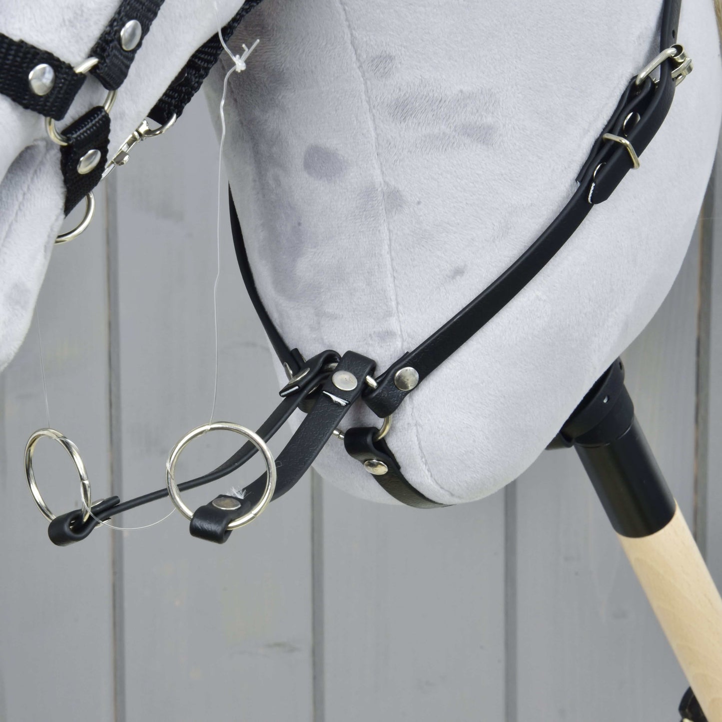 Detailní záběr hlavy plyšového Hobby Horse Martingale černý od Hobby Horse LarDen s černou uzdečkou, koženkovým martingalem a kovovými kroužky na dřevěné tyči, na pozadí šedých dřevěných panelů.