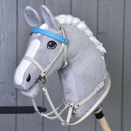 Hobby Horse LarDen se šedým plyšovým Hobby Horse se spletenou bílou hřívou, upevněným na dřevěné tyči, a s koženkovým setem Aqua – uzdečka, otěže a martingal – před šedou dřevěnou stěnou.
