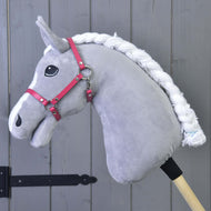 Šedý plyšový Hobby Horse LarDen s bílou spletenou hřívou, černým okem a ohlávkou malinová (vel. M), připevněný k dřevěné tyči, před šedou dřevěnou stěnou s panely.