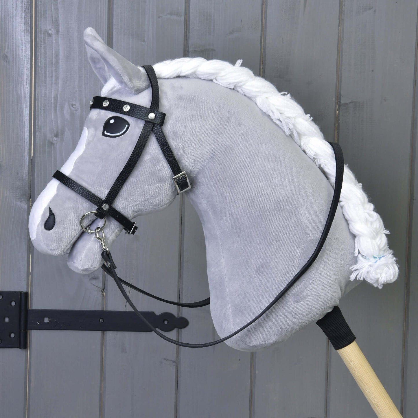 Šedý Hobby Horse s bílou hřívou, s kompletní černou uzdečkou s kamínky od Hobby Horse LarDen a odnímatelnými otěžemi, nasazený na dřevěné tyči před dřevěnou stěnou.