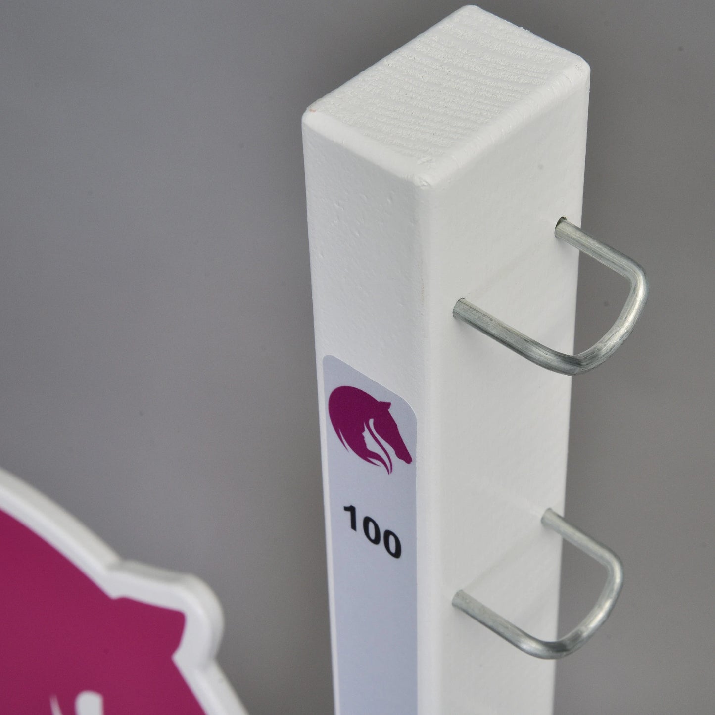 Bílý svislý stojan od Hobby Horse LarDen, označený „LarDen jump 110 cm / 43 inch“, se dvěma kovovými háčky, fialovou ikonou koňské hlavy a číslem 100, naznačuje, že je určen pro nastavitelné Hobby Horse překážky. V pozadí je růžovobílá cedule.
