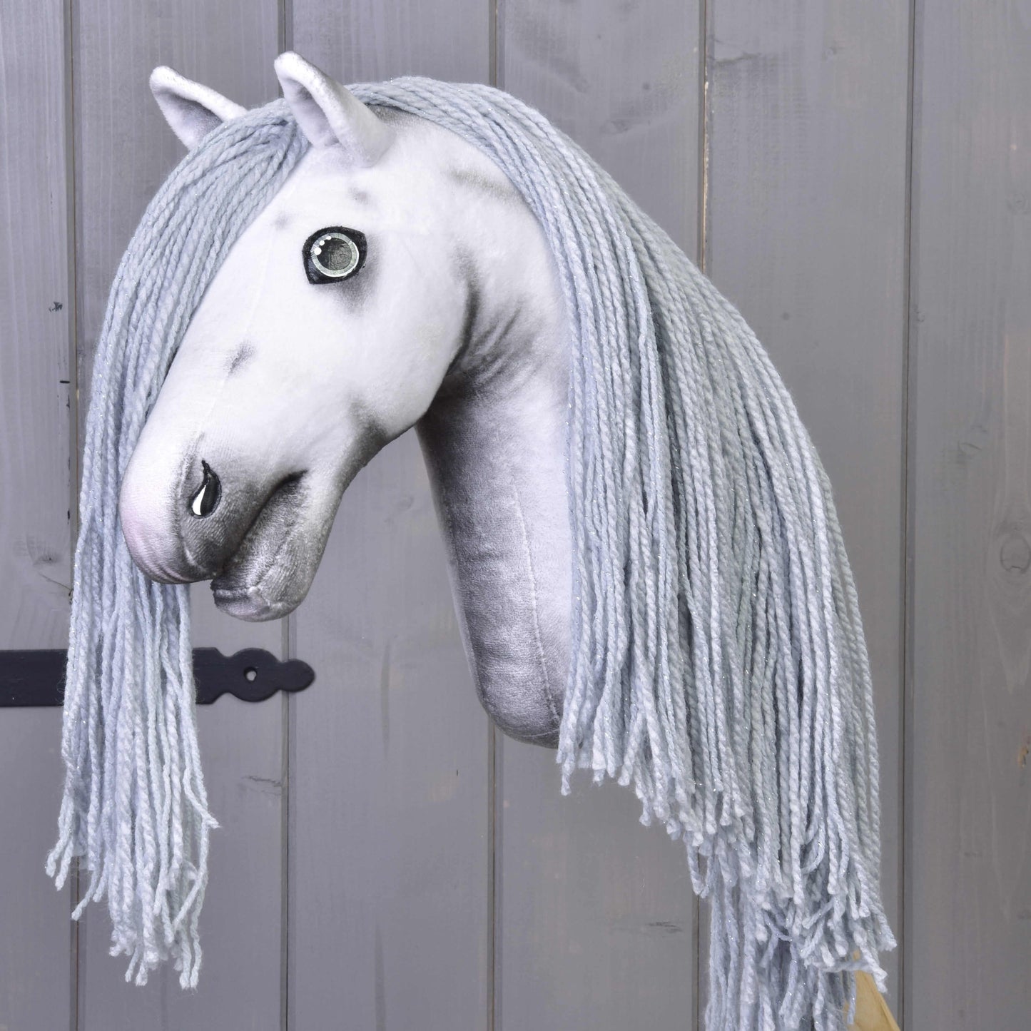 Hobby Horse jednorožec Silver s uzdou a ohlávkou od Hobby Horse LarDen s bílou plyšovou hlavou, dlouhou spletenou světle šedou hřívou z příze, nasazený na tyči a zobrazený na šedém dřevěném pozadí.