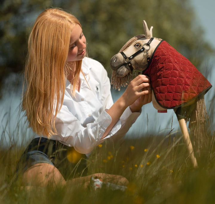 Mladý člověk s dlouhými blond vlasy a v bílém tričku sedí ve vysoké trávě a jemně upravuje uzdečku Hobby Horse s červeným prošívaným přehozem, na pozadí rozmazané zeleně, a vystihuje nápaditého ducha Hobby Horsing.