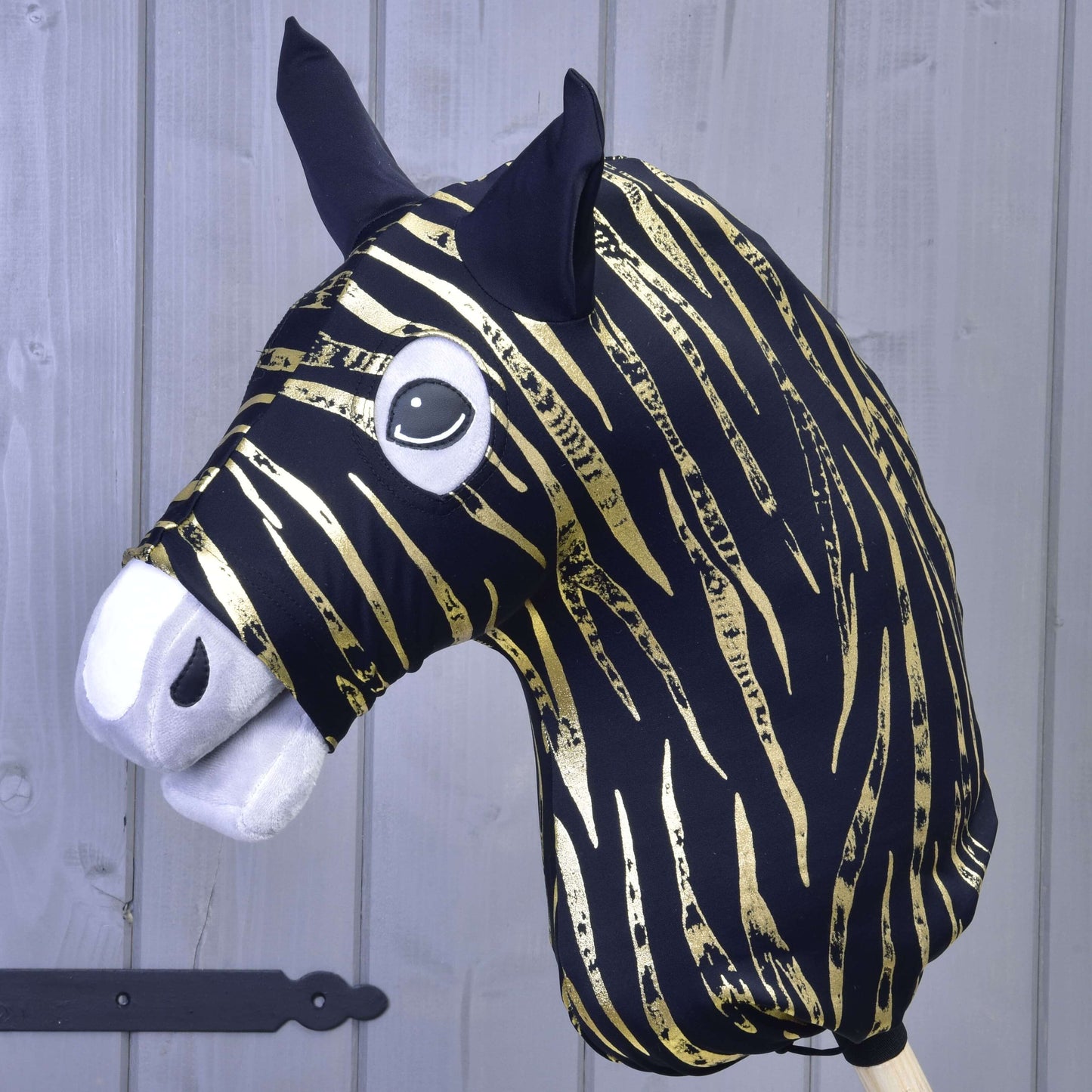 Hobby Horse LarDen ochranný oblek Zebra Gold (Lycra) s černo-zlatou pruhovanou hlavou zebry, černými ušima, bílým plstěným čenichem a velkýma kreslenýma očima, zobrazený před světle šedou dřevěnou stěnou.