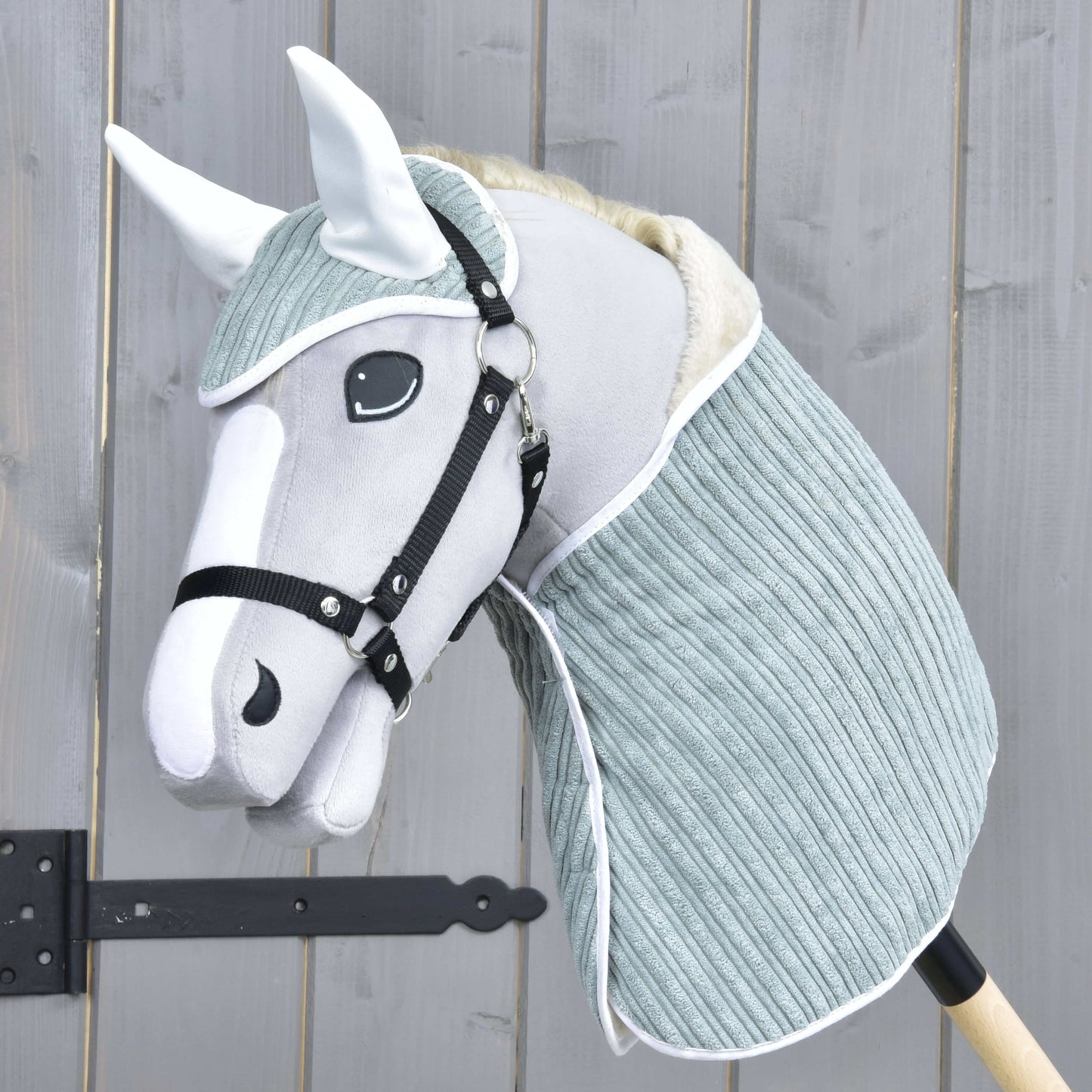 Set Manchester Little Green od Hobby Horse LarDen se šedo-bílým Hobby Horse s měkkou látkovou hlavou, černou uzdečkou, čabrakou a světle modrozeleným krytem krku na dřevěné tyči, zobrazený před dřevěným plotem.