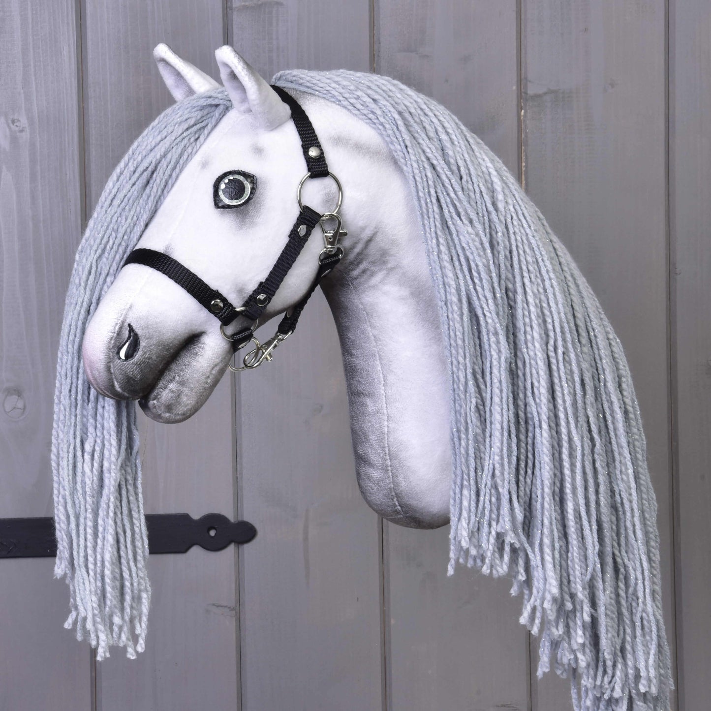 Hobby Horse jednorožec Silver s uzdou a ohlávkou od Hobby Horse LarDen s ručně vyrobenou bílou látkovou hlavou, černou uzdou a světle šedou hřívou z příze na šedém dřevěném pozadí.