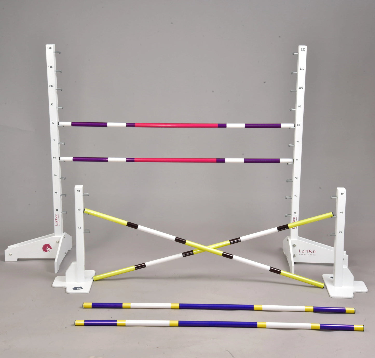 Set překážek pro Hobby Horsing od Hobby Horse LarDen s nastavitelnými bílými stojany s měřicími značkami a barevnými tyčemi ve fialové, růžové, žluté a černé barvě – ideální pro skoky nebo cavaletti, některé tyče jsou postavené a jiné leží na podlaze.