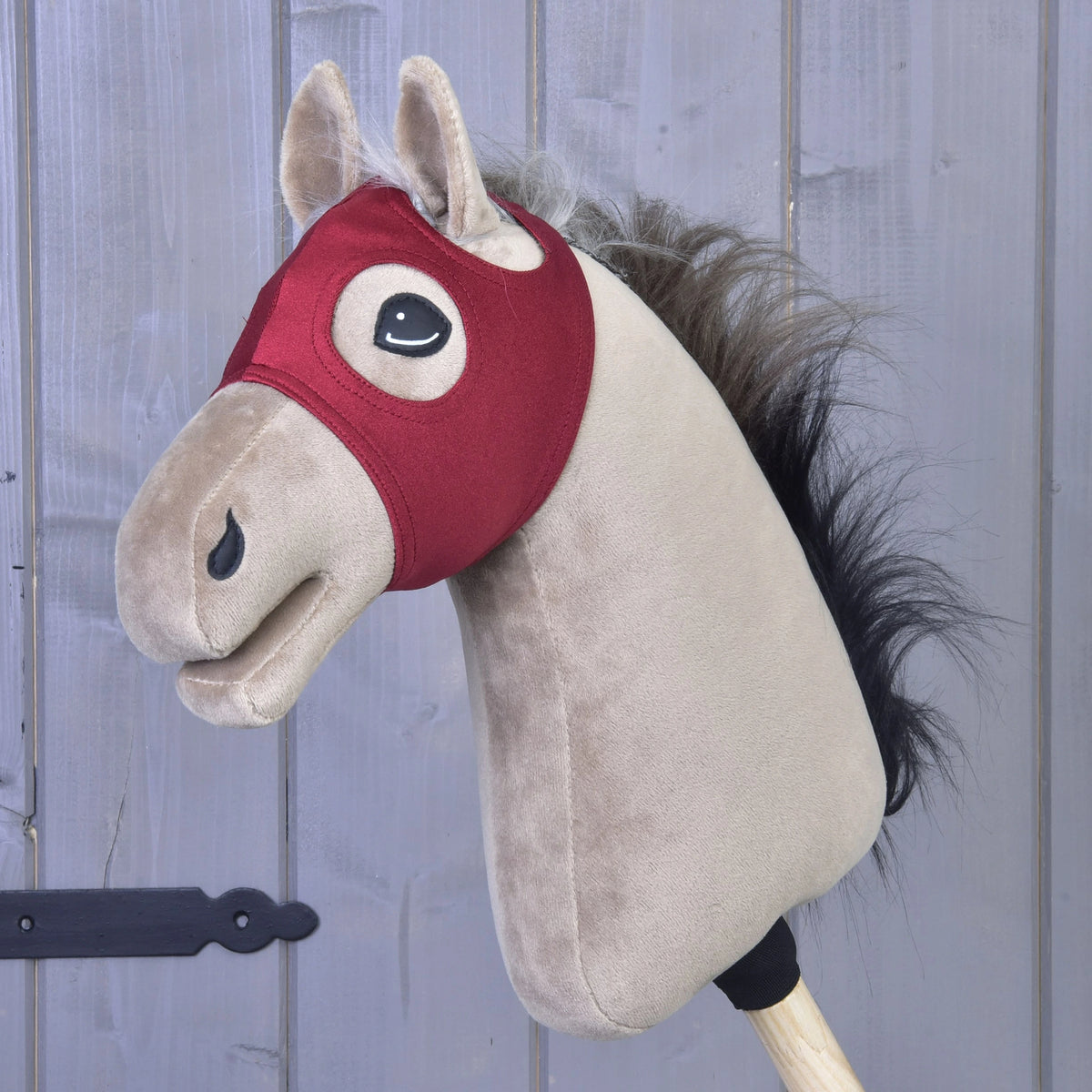 Sametová maska na koně bez uší od Hobby Horse LarDen je nasazená na dřevěné tyči před šedým dřevěným obložením a spojuje klasické kouzlo s praktickým designem; tato půvabná pratelná hračka nabízí hodiny nápadité hry a snadnou údržbu