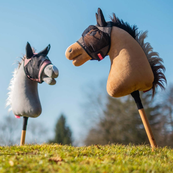 Dva hračkoví hobby horse s plyšovými hlavami, jeden šedý a jeden hnědý, stojí vzpřímeně na travnaté zemi proti jasně modré obloze a rozmazaným stromům. Oba koně mají malé látkové uzdečky, připravené na nápadité dobrodružství v Hobby Horsing.