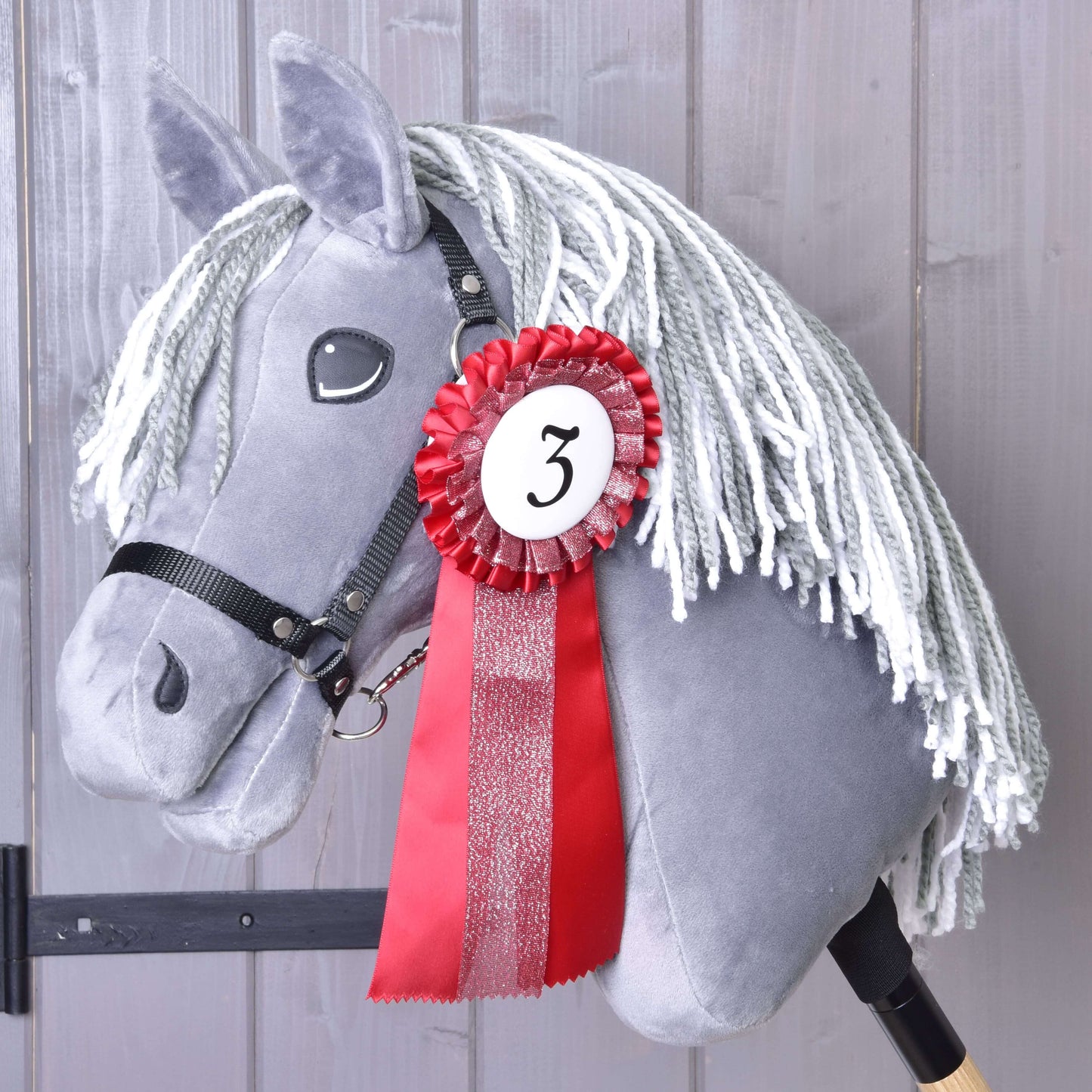 Šedý plyšový Hobby Horse s bílou přízovou hřívou, černou uzdečkou a červeno-bílou Hobby Horse LarDen „Set Hobby Horse Kokarda NEW“ připnutou s číslem 3 stojí před šedými dřevěnými panely.