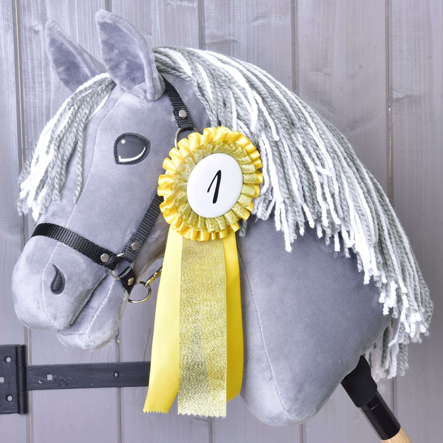 Set Hobby Horse Kokarda NEW od Hobby Horse LarDen s žlutou kokardou za první místo označenou „1“ na šedém Hobby Horse s bílou přízovou hřívou, zobrazený na pozadí dřevěné stěny.