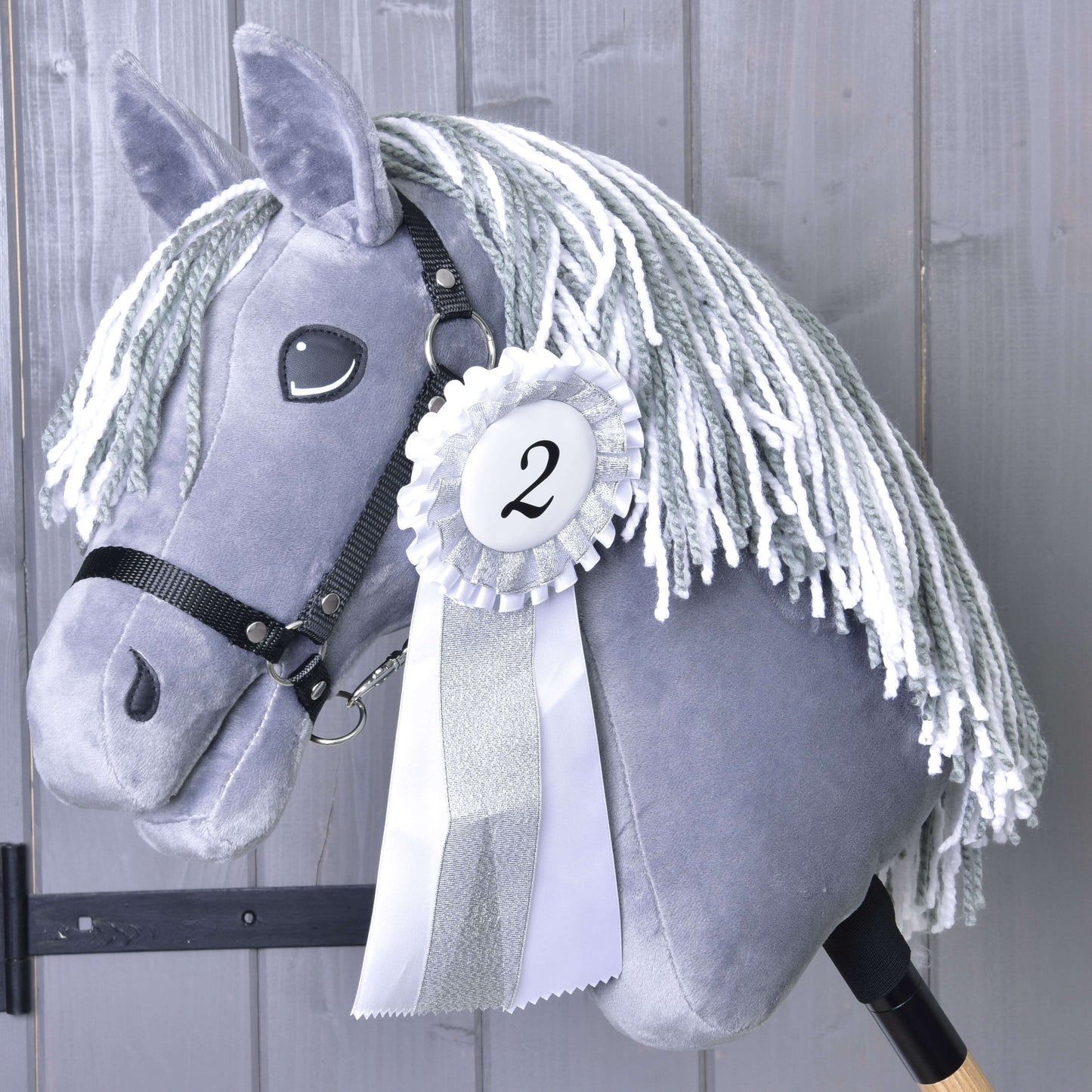 Šedý Hobby Horse s bílou přízovou hřívou a černou uzdečkou na dřevěné tyči má Set Hobby Horse Kokarda NEW (druhé místo, stříbrné lemování, číslo 2) od Hobby Horse LarDen na pozadí šedých dřevěných prken.