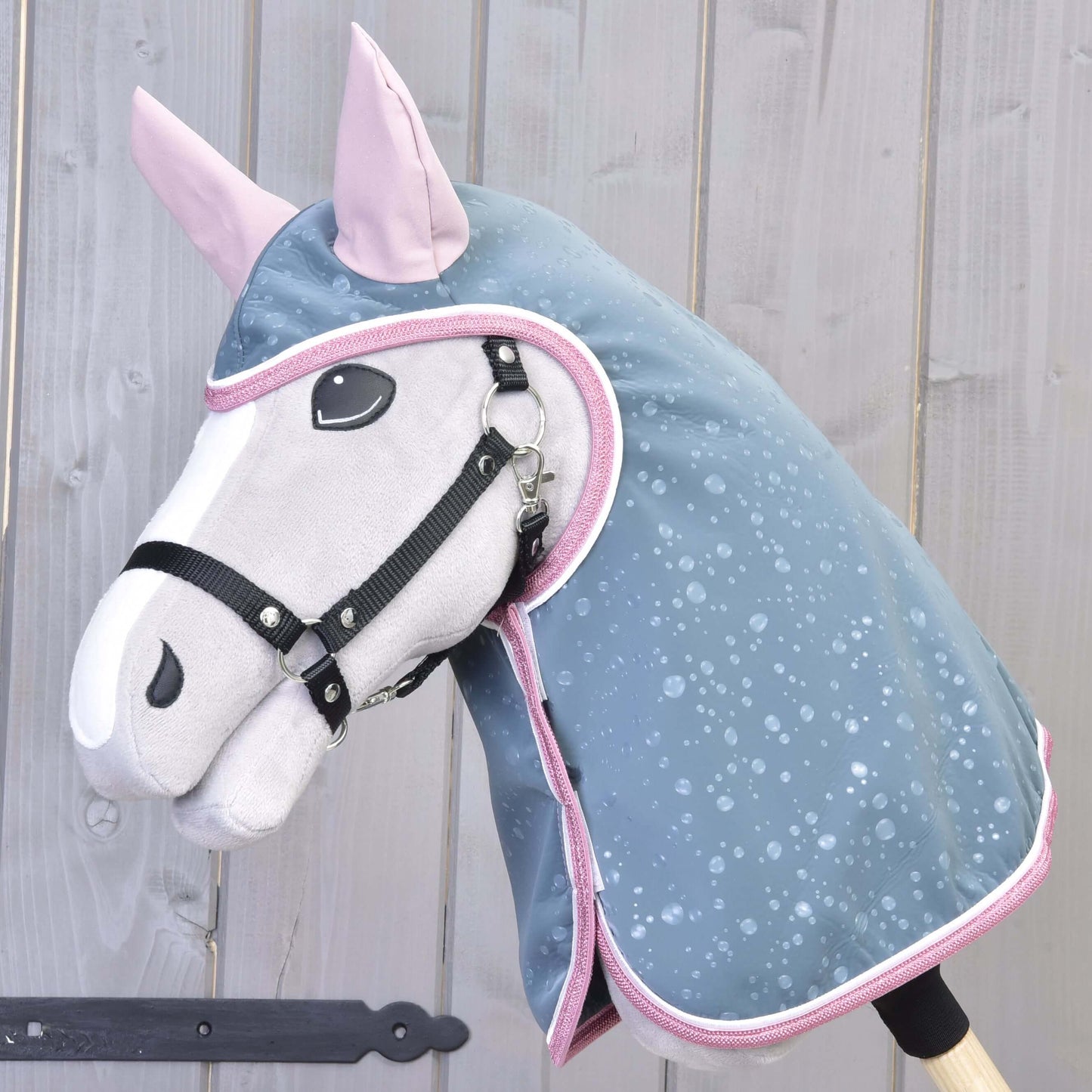 Hobby Horse LarDen „Raincoat Best“ má plyšovou koňskou hlavu s černou uzdečkou a modrou pláštěnkou do každého počasí s růžovým lemováním, nasazenou na dřevěné tyči před stěnou ze světlého dřeva.