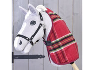 Šedá hračka Hobby Horse od Hobby Horse LarDen, s dekou Deka stájová Christmas v červené kostce a černou uzdečkou, je nasazená na dřevěné tyči a umístěná před pozadím ze světlých dřevěných panelů.