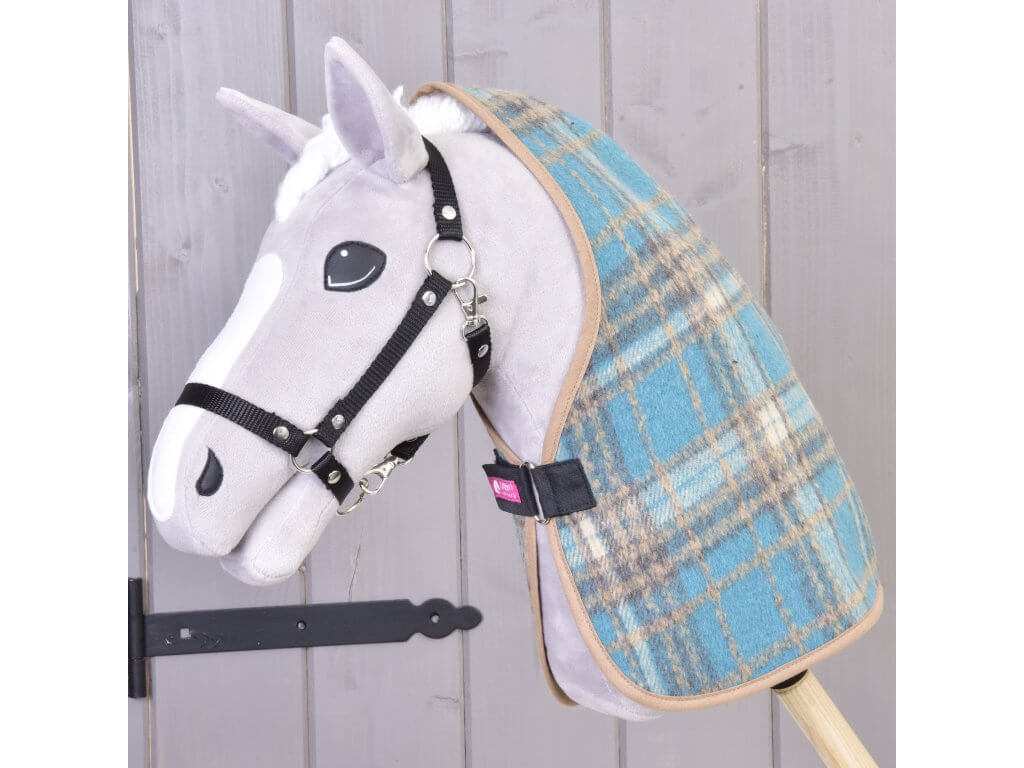 Plyšový Hobby Horse od Hobby Horse LarDen se šedo-bílým obličejem, černou uzdečkou a dekou Deka stájová Scottish Cube v modré kostce stojí na tyči před světlou dřevěnou stěnou.