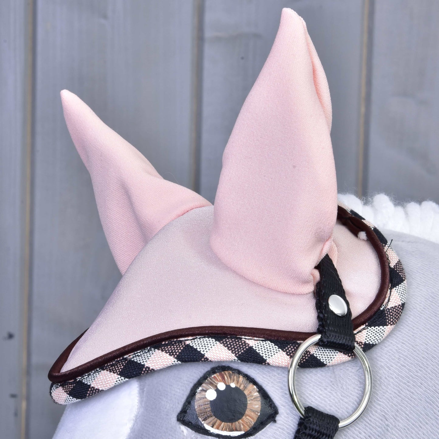 Detail plyšové hračky Set Abby od Hobby Horse LarDen s růžovou čabrakou na uši s kostkovaným lemováním a černým páskem. Na dřevěném pozadí jsou vidět velké látkové uši a prošité oko.