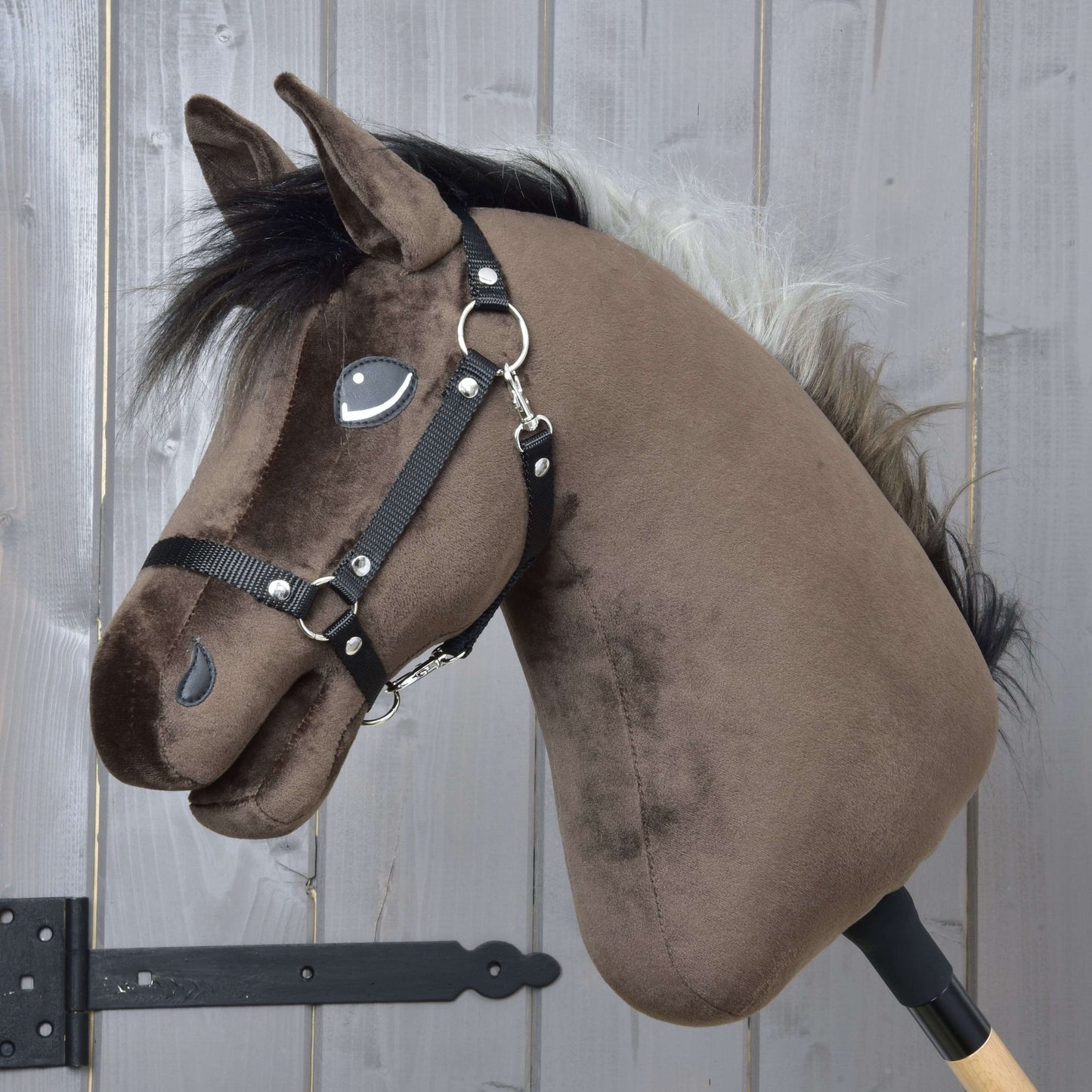 Hobby Horse Alfredo s černou ohlávkou od Hobby Horse LarDen má hnědou plyšovou hlavu ve stylu plnokrevníka, černou hřívu, bílou ofinu a černou ohlávku, je nasazený na dřevěné tyči a umístěný před šedým dřevěným pozadím.