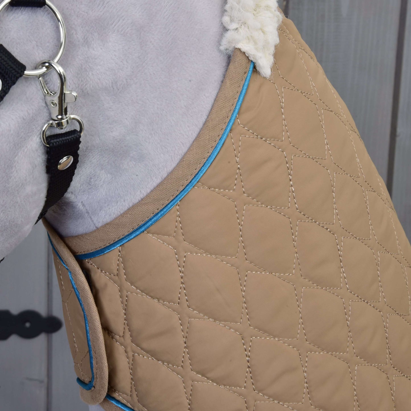 Detailní záběr na psí kabátek Set Almond Star od Hobby Horse LarDen s béžovou prošívanou látkou s modrým lemováním, huňatou bílou podšívkou a černým popruhem s kovovou přezkou na dřevěném pozadí.